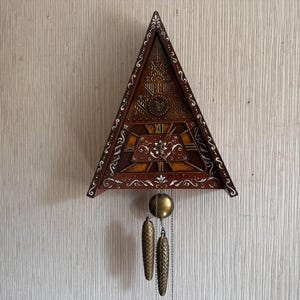 May include: A triangular wooden wall clock featuring a dark brown finish with white painted scrollwork details. The clock face displays Roman numerals and a central floral motif. It includes a brass pendulum and two hanging pinecone-shaped weights.