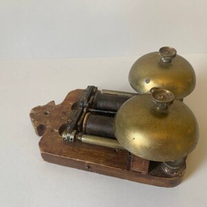 May include: Antique double bell door chime with a wooden base and brass bells. The chime has two brass bells with a silver knob on top of each bell. The bells are attached to a wooden base with a mechanism for ringing the bells.