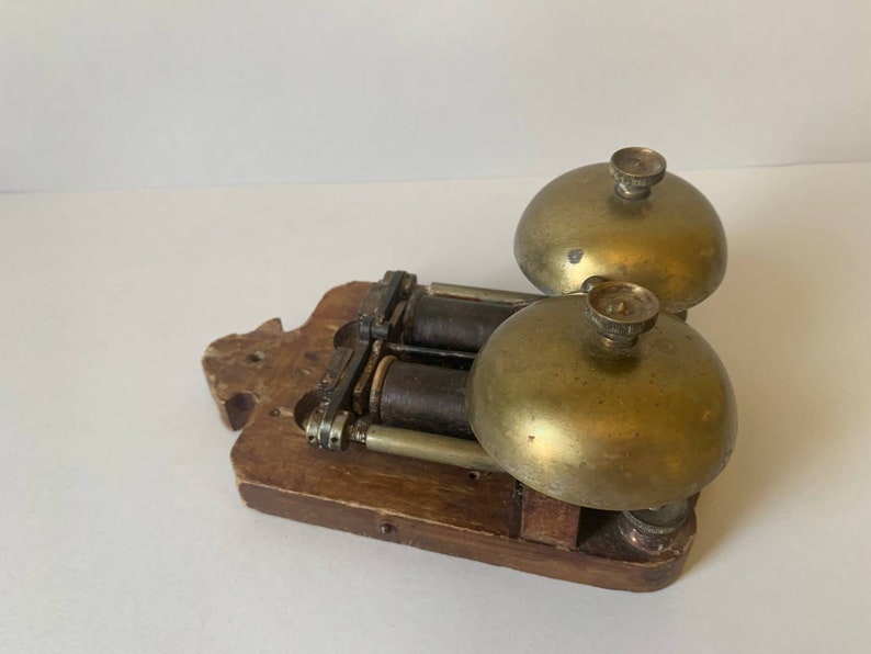 May include: Antique double bell door chime with a wooden base and brass bells. The chime has two brass bells with a silver knob on top of each bell. The bells are attached to a wooden base with a mechanism for ringing the bells.