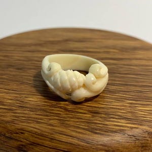 May include: Ivory-coloured ring with a detailed, carved design. The ring features a central sculpted element with swirling patterns and rounded shapes. The ring is displayed on a wooden surface.