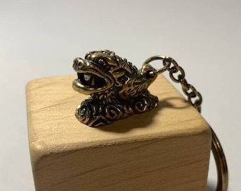 Puede incluir: Un llavero de metal dorado con un diseño de dragón detallado. El llavero está unido a un bloque de madera.