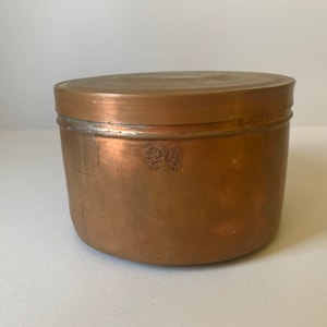 May include: A copper storage container with a lid. The container has a circular shape and a smooth, shiny surface. The lid has a slightly raised edge.