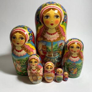 May include: A set of seven Russian nesting dolls, or Matryoshka dolls, in various sizes. Each doll features a painted face, floral headscarf, and detailed clothing. The largest doll is approximately 7 inches tall.