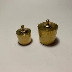 Op de afbeelding: Twee messing gewichten met een knop aan de bovenkant. Het grotere gewicht is ongeveer 5 cm hoog en het kleinere ongeveer 2,5 cm hoog.