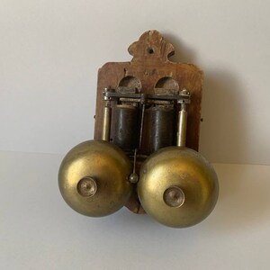 May include: Antique wooden wall mounted doorbell with two brass bells. The doorbell has a wooden frame and two brass bells.