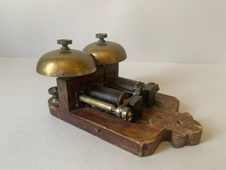May include: Antique brass double bell door chime with a wooden base. The chime has two brass bells and a wooden base with a decorative design.