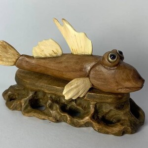 May include: A handcrafted wooden fish sculpture with large, expressive eyes and fins. The fish is a warm brown color with lighter beige fins and is mounted on a carved wooden base. The sculpture is a unique decorative item.