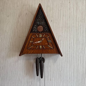 May include: A triangular wooden wall clock featuring a dark brown carved upper section with a circular emblem and a lower section with a trapezoidal face. The clock has gold-colored hands, raised gold-colored hour markers, and two hanging pinecone weights.