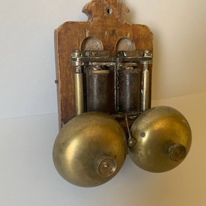 May include: Antique wooden wall mounted doorbell with two brass bells. The doorbell has a wooden frame and two brass bells. The bells are attached to a metal mechanism with two coils.