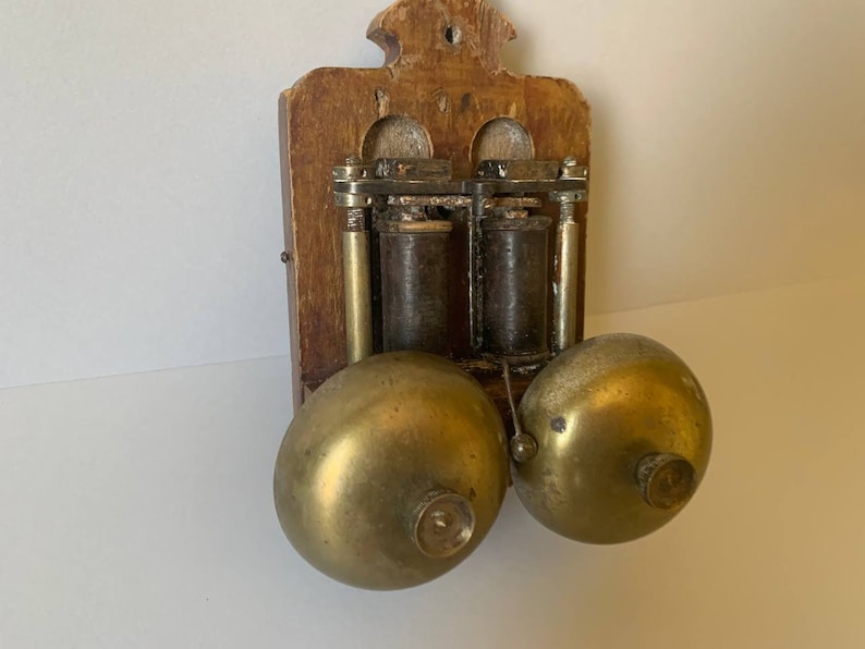 May include: Antique wooden wall mounted doorbell with two brass bells. The doorbell has a wooden frame and two brass bells. The bells are attached to a metal mechanism with two coils.
