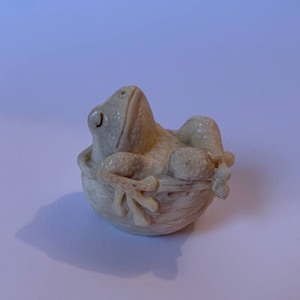 May include: A white ceramic frog figurine sitting in a small, white ceramic bowl. The frog is looking up and has a detailed, textured surface.