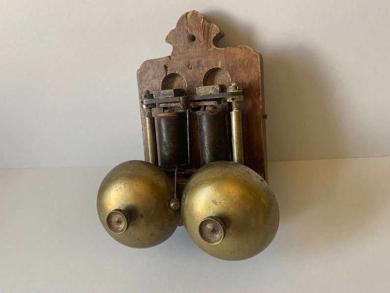 May include: Antique wooden wall mounted doorbell with two brass bells. The doorbell has a wooden frame with a decorative top and two brass bells.