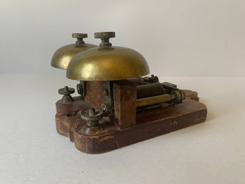May include: Antique brass double bell door chime with a wooden base. The chime has two brass bells with a mechanism in the center.