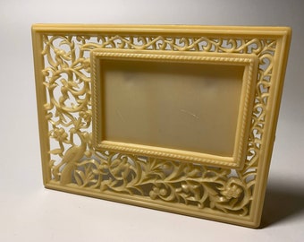 May include: A cream-colored plastic picture frame with an intricate, carved design featuring a peacock and floral motifs.