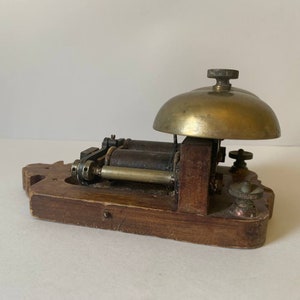 May include: Antique wooden doorbell with two brass bells and a metal mechanism. The doorbell is mounted on a wooden base.