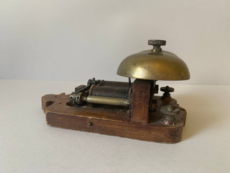 May include: Antique wooden doorbell with two brass bells and a metal mechanism. The doorbell is mounted on a wooden base.