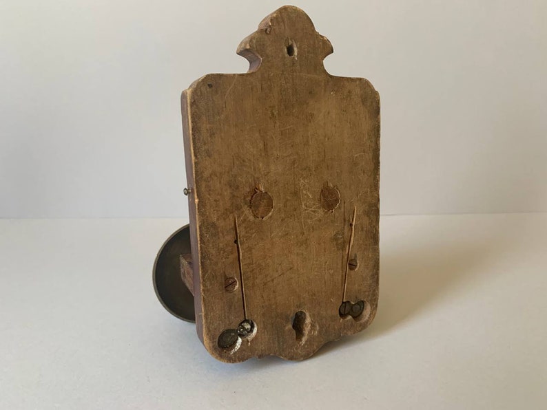 May include: A wooden wall mount with a decorative design and a metal ring on the bottom. The back of the mount is visible, showing the wood grain and a few holes.