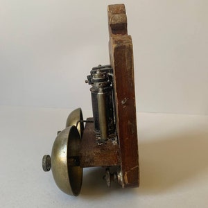 May include: A vintage wooden doorbell with two brass bells. The doorbell has a black metal mechanism and is mounted on a wooden plaque.