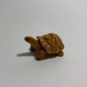 Op de afbeelding: Een klein, gedetailleerd beeldje van een schildpad. De schildpad is bruin met een getextureerd schild en zichtbare poten en kop. Het beeldje staat tegen een effen witte achtergrond.