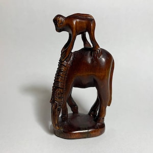 May include: A dark brown wooden sculpture of a monkey riding a horse. The monkey is sitting on the horse's back and has its arms around the horse's neck. The horse is standing on all four legs and has its head down.