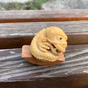 May include: A small, intricately carved ivory dragon figurine, curled up on a wooden base. The dragon has a detailed face with a small black eye and is a light cream color.