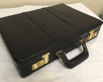 May include: A black leather briefcase with a handle and gold-colored combination locks. The briefcase has three sections on the top and is closed. The briefcase is on a white surface.
