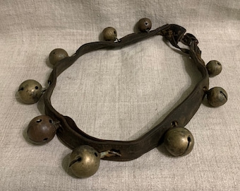 May include: A brown leather strap with eight brass bells attached. The strap is worn and has a rustic look.