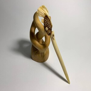 May include: A carved wooden hair stick featuring a fish design. The fish is brown with a detailed scale pattern and a small eye. The hair stick is light beige and has a small floral design. The base is a light brown, intertwined shape.