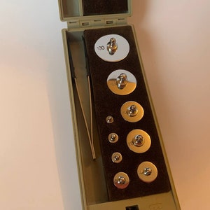 Op de afbeelding: Een set verchroomde gewichten in een groene koffer. De gewichten zijn in verschillende maten, van 1 gram tot 100 gram. De koffer heeft een zwarte binnenkant met een fluwelen voering.