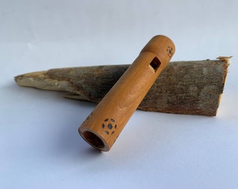 Puede incluir: Una flauta de madera con un acabado marrón y diseños geométricos negros. La flauta está apoyada en un trozo de madera.