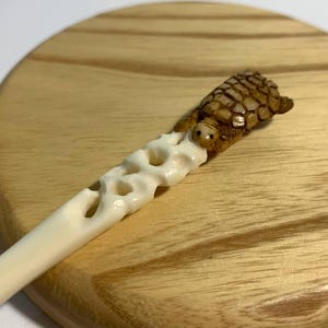 May include: A carved wooden hair stick featuring a detailed turtle figurine. The turtle is brown and tan, perched on a cream-colored stick with decorative cutouts. The stick rests on a wooden surface.