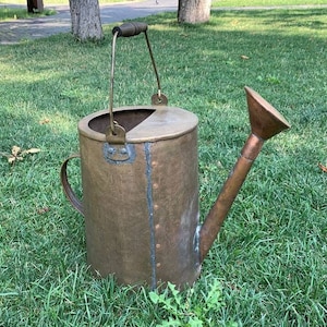 May include: A vintage copper watering can with a wooden handle and a spout. The can has a worn, rustic look and is sitting on a bed of green grass.