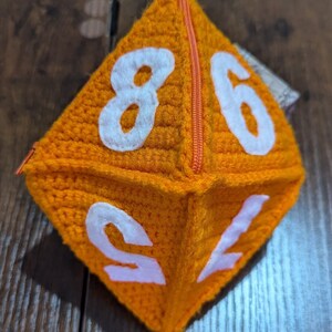 May include: Orange crocheted dice pouch with a zipper closure and white numbers 8, 6, 1, and 2.