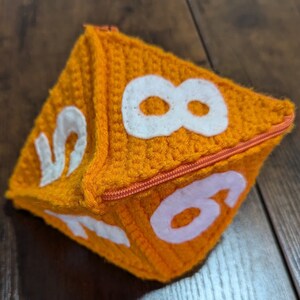 May include: A crocheted orange dice with white numbers 2, 6, and 8. The dice has a zipper closure.
