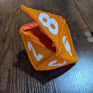 Könnte beinhalten: Orangefarbene gehäkelte Würfeltasche mit Reißverschlussöffnung. Die Tasche ist würfelförmig und hat weiße Zahlen auf jeder Seite.
