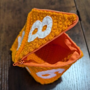 May include: Orange crocheted pouch with a zipper closure and white felt numbers 8 and 8 on the front.