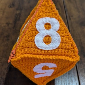 May include: Orange crocheted dice with white numbers 8 and 9. The dice has a zipper on one side.