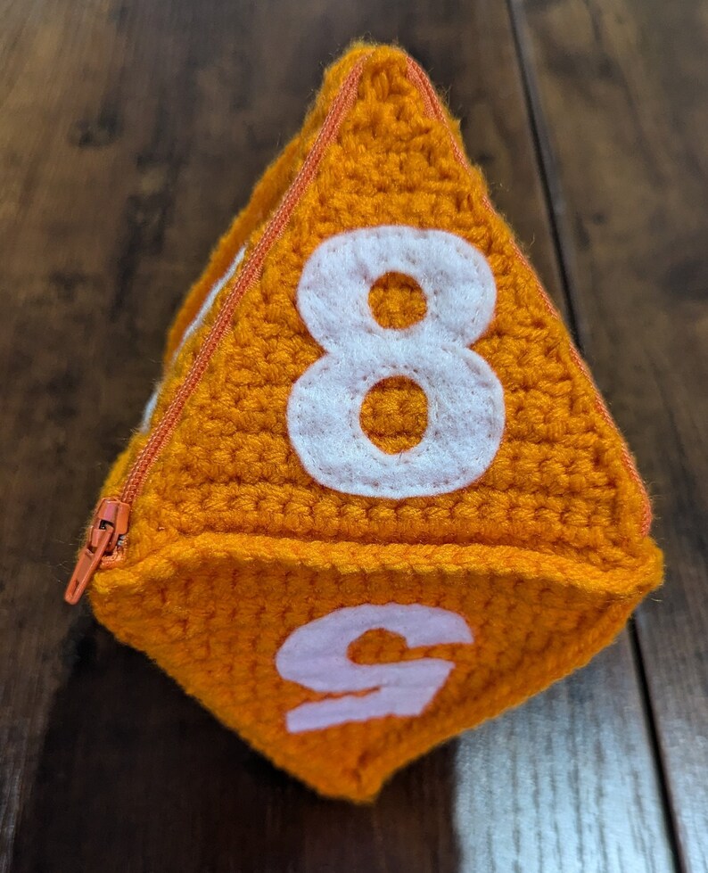 May include: Orange crocheted dice with white numbers 8 and 9. The dice has a zipper on one side.