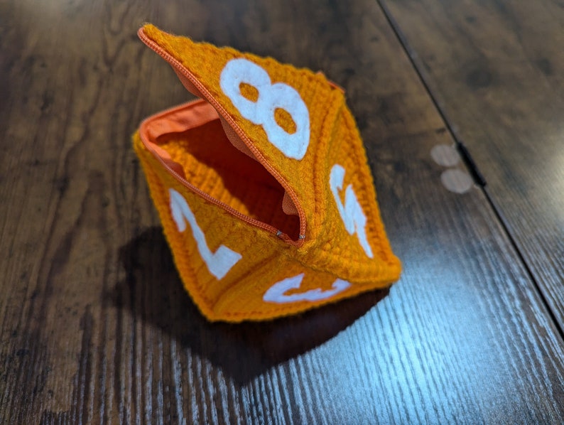 May include: Orange crocheted dice pouch with a zipper opening. The pouch is shaped like a dice and has white numbers on each side.