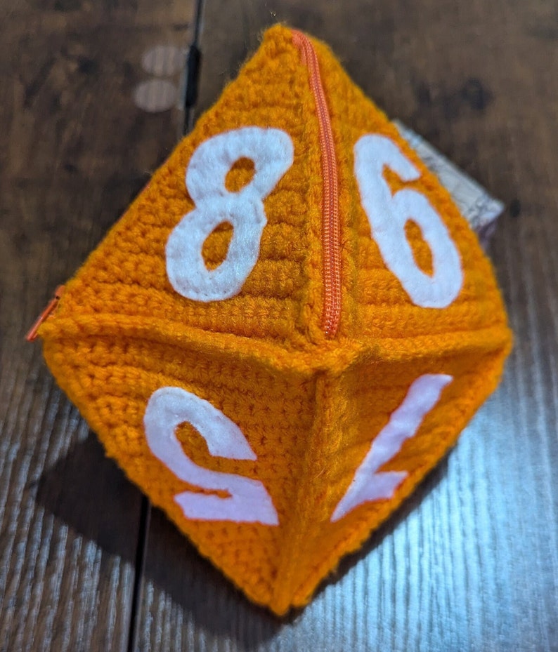 May include: Orange crocheted dice pouch with a zipper closure and white numbers 8, 6, 1, and 2.