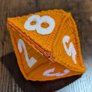 May include: A crocheted orange dice with white numbers 2, 3, 4, 5, 6, 7, and 8. The dice has a zipper closure.