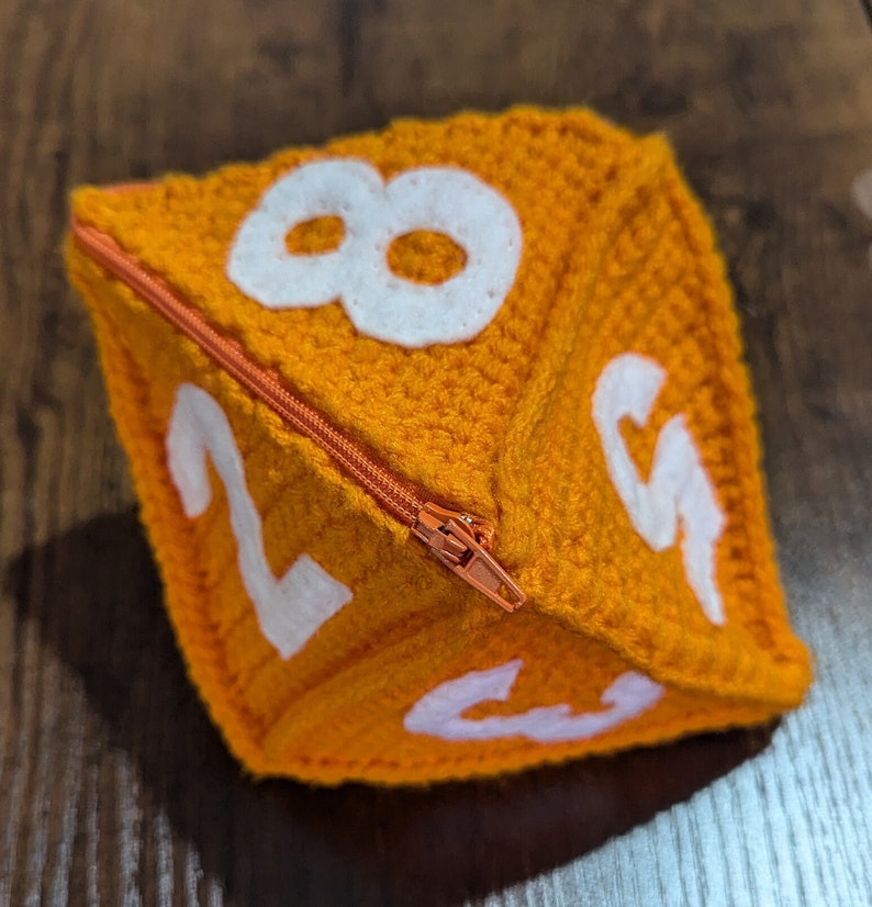 May include: A crocheted orange dice with white numbers 2, 3, 4, 5, 6, 7, and 8. The dice has a zipper closure.