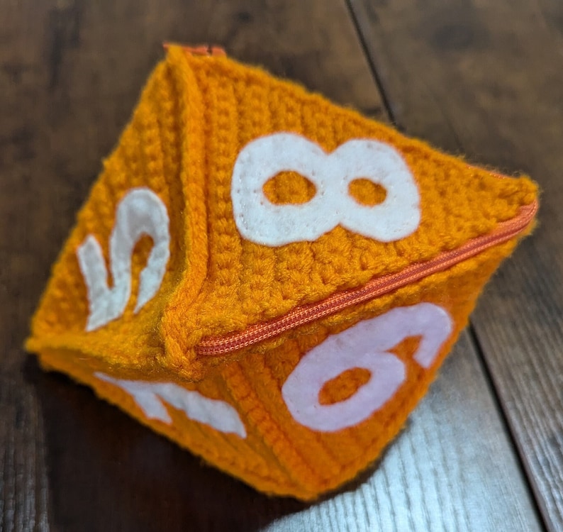 May include: A crocheted orange dice with white numbers 2, 6, and 8. The dice has a zipper closure.