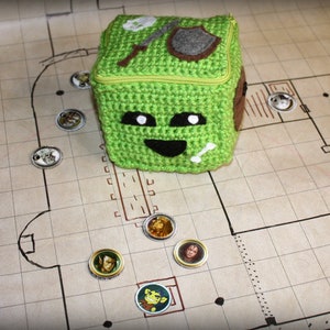 May include: A green crocheted dice bag with a smiling face, white eyes, and a bone sticking out of its mouth. The bag is sitting on a brown and white gridded surface with several small round tokens with images of people on them.