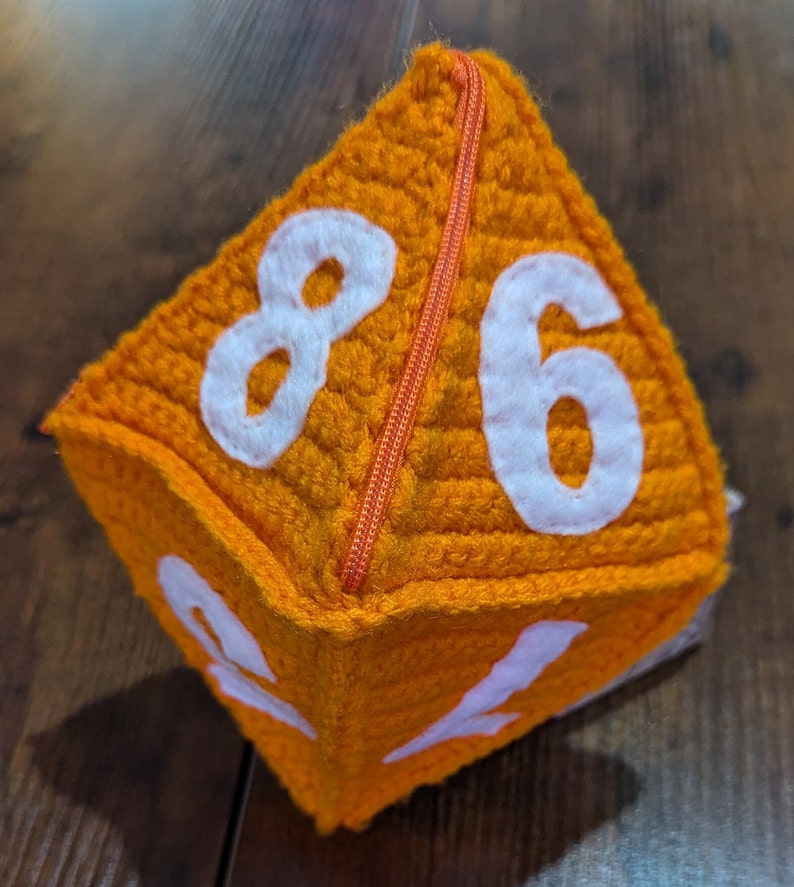 May include: A plush orange dice with white numbers 8, 6, 1, and 2. The dice has a zipper sewn down the middle.