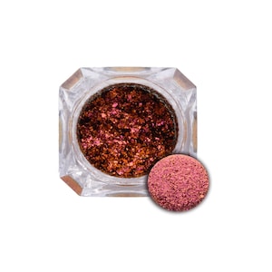 May include: A clear, faceted jar filled with shimmering, multi-toned glitter. The glitter appears to be a mix of bronze, copper, and pink hues. A small, round swatch of the glitter is displayed next to the jar, showcasing the color.