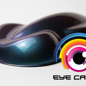 May include: A close-up of a dark purple and teal iridescent car paint sample. The paint is very shiny and has a metallic finish. The sample is sitting on a white surface. The Eye Candy logo is visible in the lower right corner of the image.