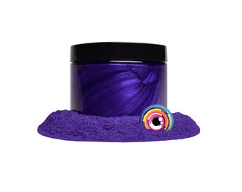 May include: A cylindrical jar with a black lid containing a deep purple, metallic, swirled substance. The jar sits in a pile of vibrant purple powder. A small, circular logo featuring a stylized eye with a rainbow-colored iris is visible.
