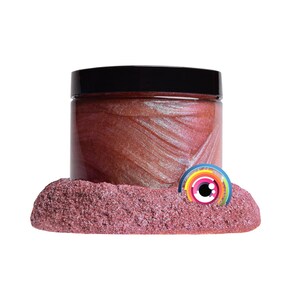 May include: A jar of shimmering, reddish-pink cosmetic product with a black lid. The jar sits on a bed of textured, pink material. A colorful, circular logo is partially visible.
