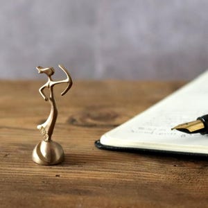 Op de afbeelding: Een gouden metalen beeldje van een persoon die een boog en pijl vasthoudt. Het beeldje staat op een ronde basis.